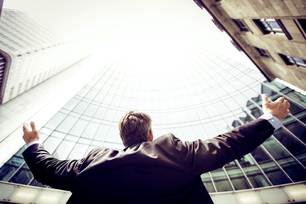 A man in a suit is facing a skyscraper. is arms are open and he is facing the white sky in an optimistic posture.