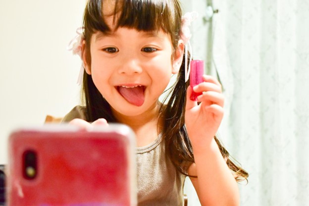 A Japanese child is holding a phone. She is sticking her tongue out looking both amused and concentrated.